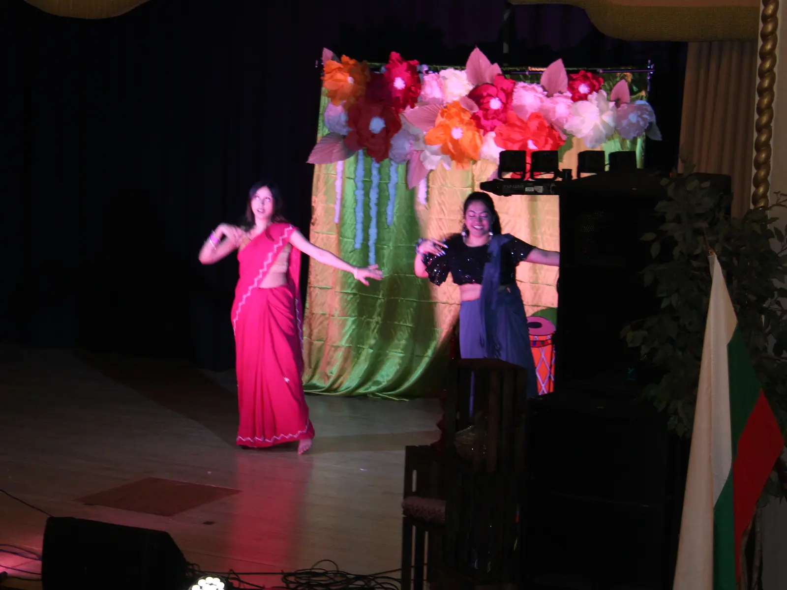 Two dancers in saris performing on stage in front of a paper flower backdrop at Boshontoboron 2025.