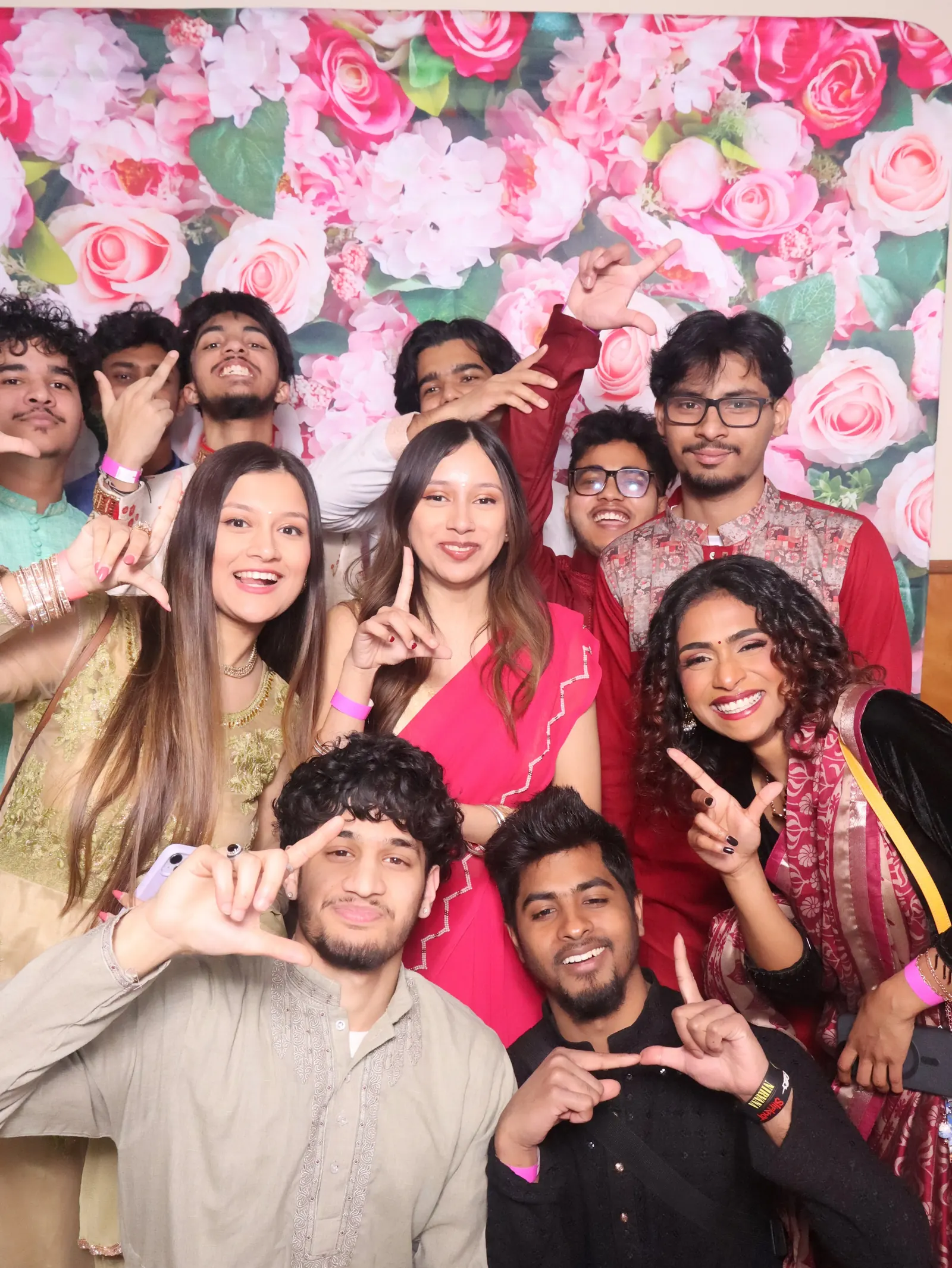 Members in saris and panjabis posing in front of a wall of pink flowers.